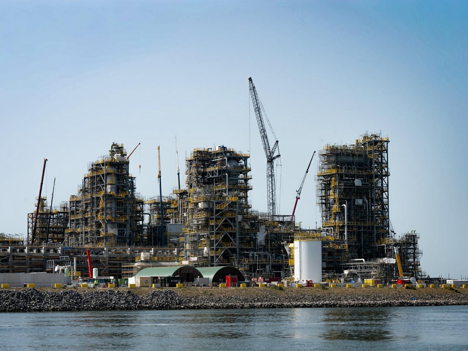 Large industrial process plant under construction beside water with cranes in place